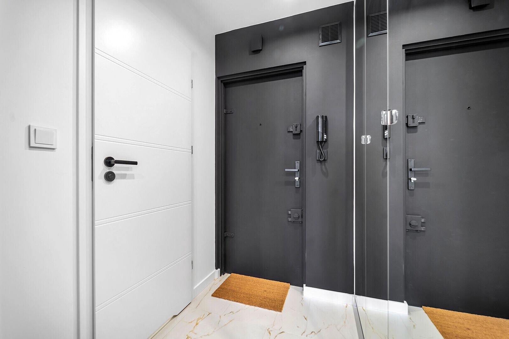 Un pasillo con una puerta blanca a la izquierda y dos puertas grises oscuras a la derecha, cada una con un teléfono y una cerradura.