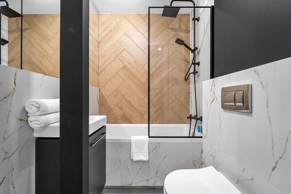 A modern bathroom with a herringbone wood-patterned wall, a glass-enclosed shower, a white sink, and a marble-tiled floor.