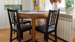Uma sala de jantar com uma mesa redonda de madeira, duas cadeiras pretas, uma cortina com padrão hexagonal, uma janela com vista para o verde e uma planta em vaso no parapeito.