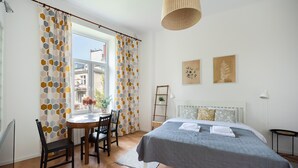 Um quarto com cama, escrivaninha, cadeiras, janela com cortinas e duas obras de arte na parede.