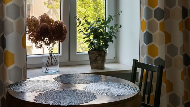 Quarto com janela, planta em vaso, um arranjo de flores secas e mesa com toalha rendada.