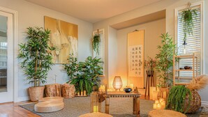A cozy living room with a wooden floor, wicker ottomans, a table with candles, and various potted plants.