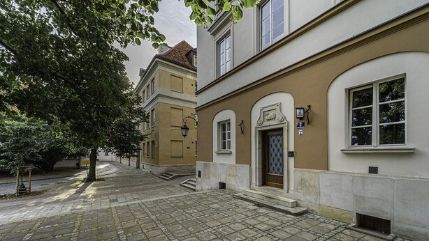 Une rue pavée, avec un bâtiment dont la porte est surmontée d’une grille en métal, un lampadaire et une enseigne indiquant le numéro 3.