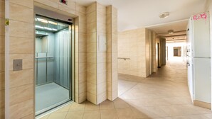 Un pasillo moderno con un ascensor de vidrio, paredes con azulejos color beige y un refrigerador blanco.