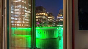 A view from a hotel room window at night, featuring a pool with green lighting, a cityscape with illuminated buildings, and a modern building with balconies.