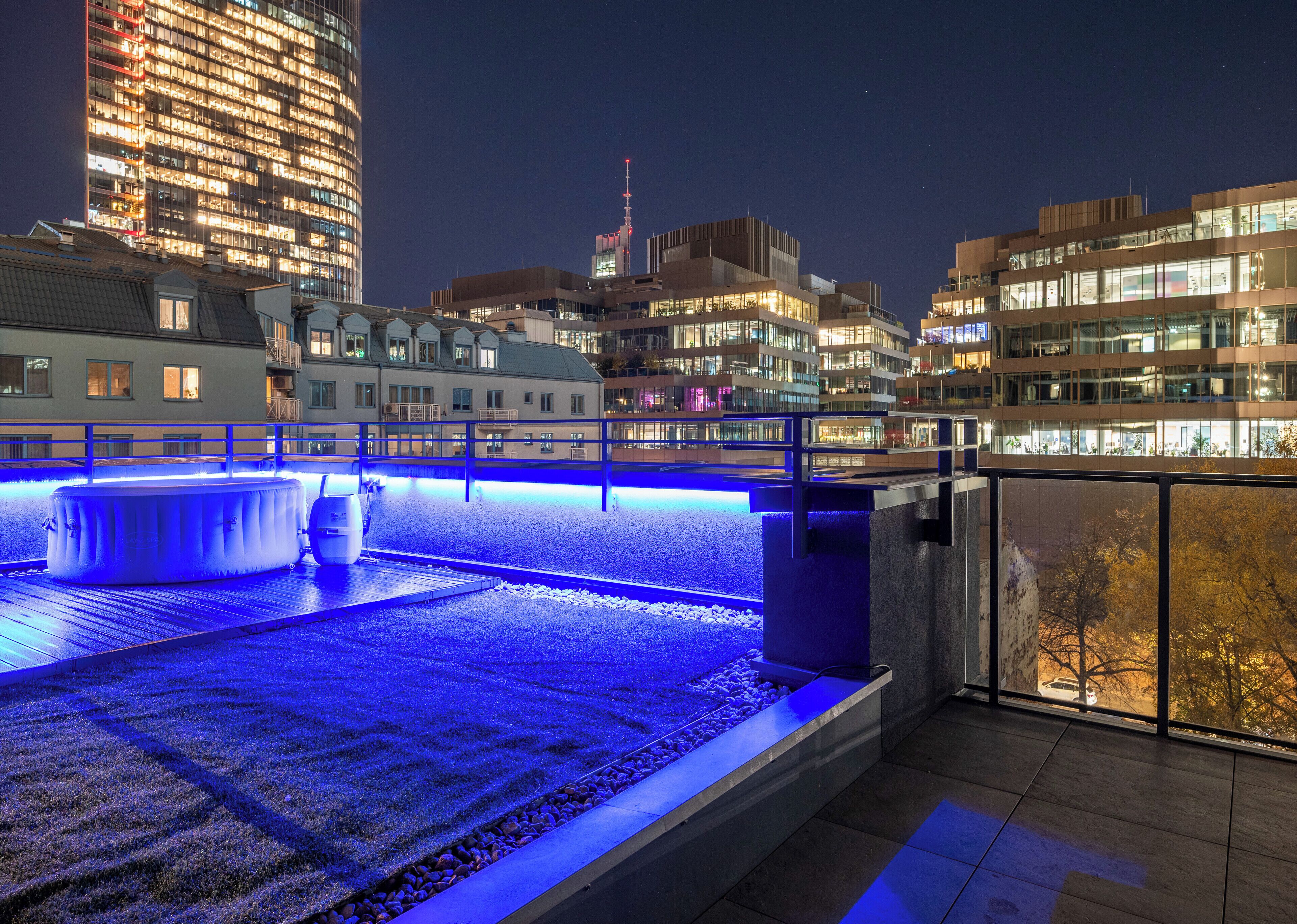 Ein Pool auf dem Dach mit blauer Beleuchtung, umgeben von modernen Gebäuden und einer nächtlichen Stadtansicht.