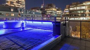 A rooftop pool with blue lighting, surrounded by modern buildings and a cityscape at night.