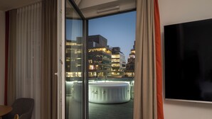 A modern hotel room with a large window, a flat-screen TV, a round hot tub, and a dining area with a table and chairs.