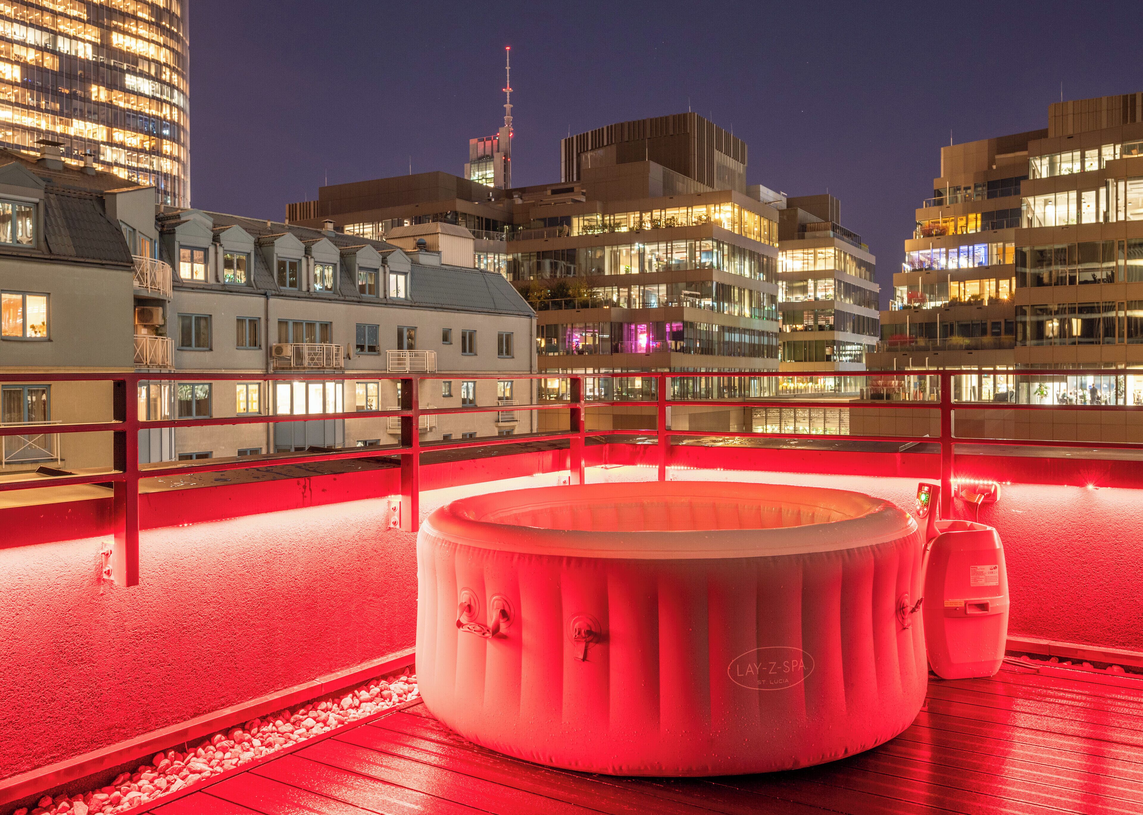 Area benessere sul tetto con vasca idromassaggio, circondata da una ringhiera illuminata di rosso, con vista sulla città di notte.