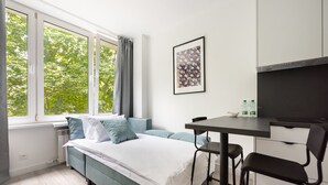 A modern bedroom with a bed, a desk, and a chair. There is a window with curtains, a radiator, and a framed picture on the wall.