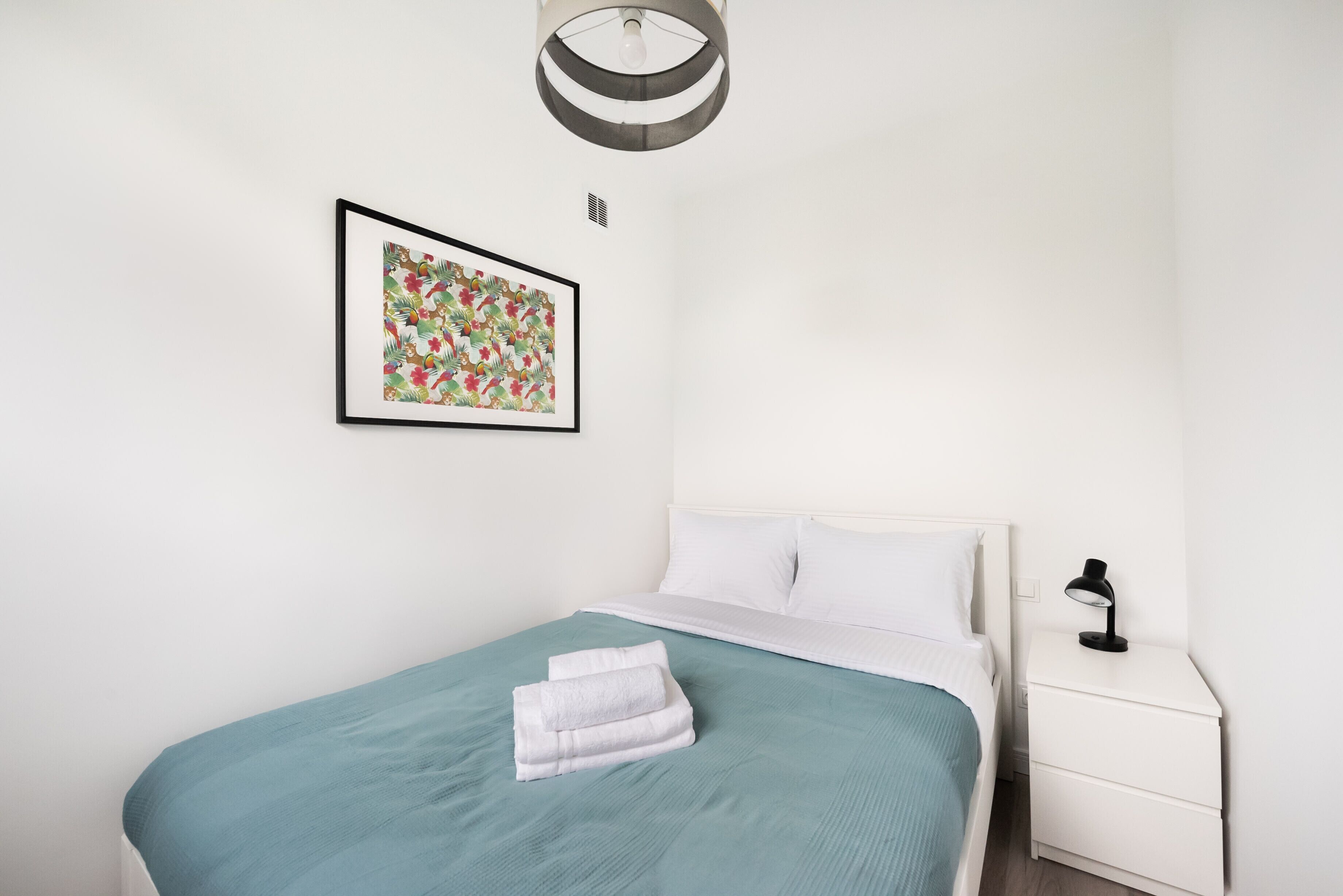 A neatly made bed with a teal bedspread and white pillows, a framed floral artwork on the wall, a bedside lamp, and a white nightstand.