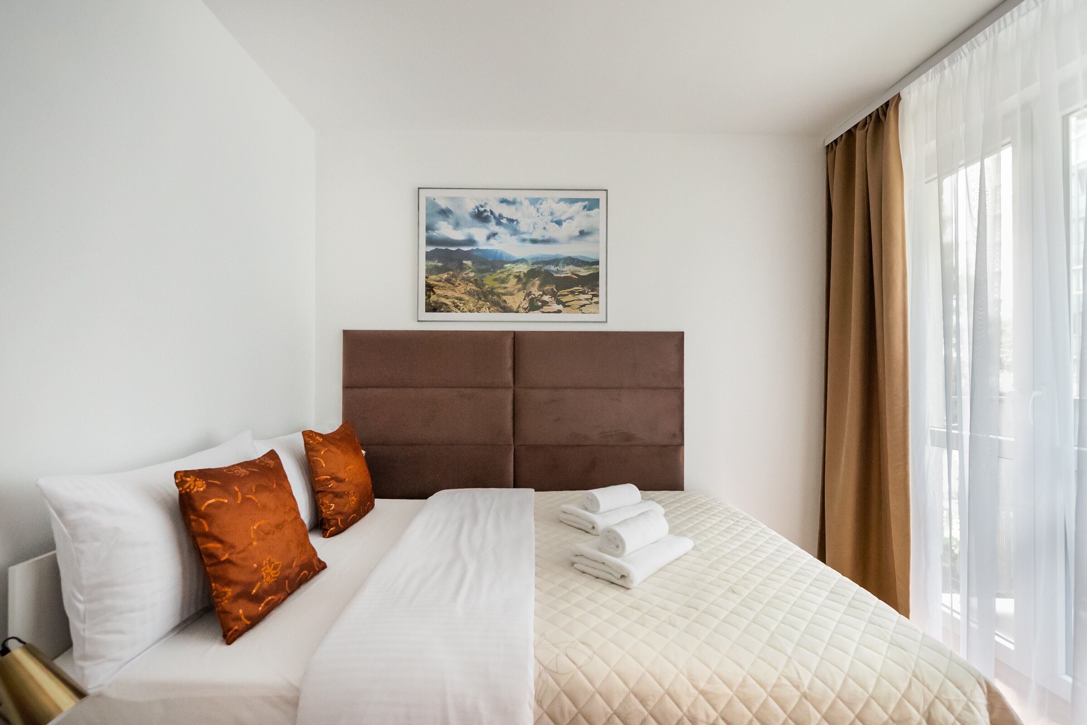 Une chambre d’hôtel avec un grand lit, une tête de lit, un tableau représentant un paysage encadré et une fenêtre avec des rideaux.