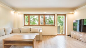 Ein modernes Wohnzimmer mit einer Couch, einem Couchtisch, einem Fernseher und großen Fenstern mit Blick auf Bäume.
