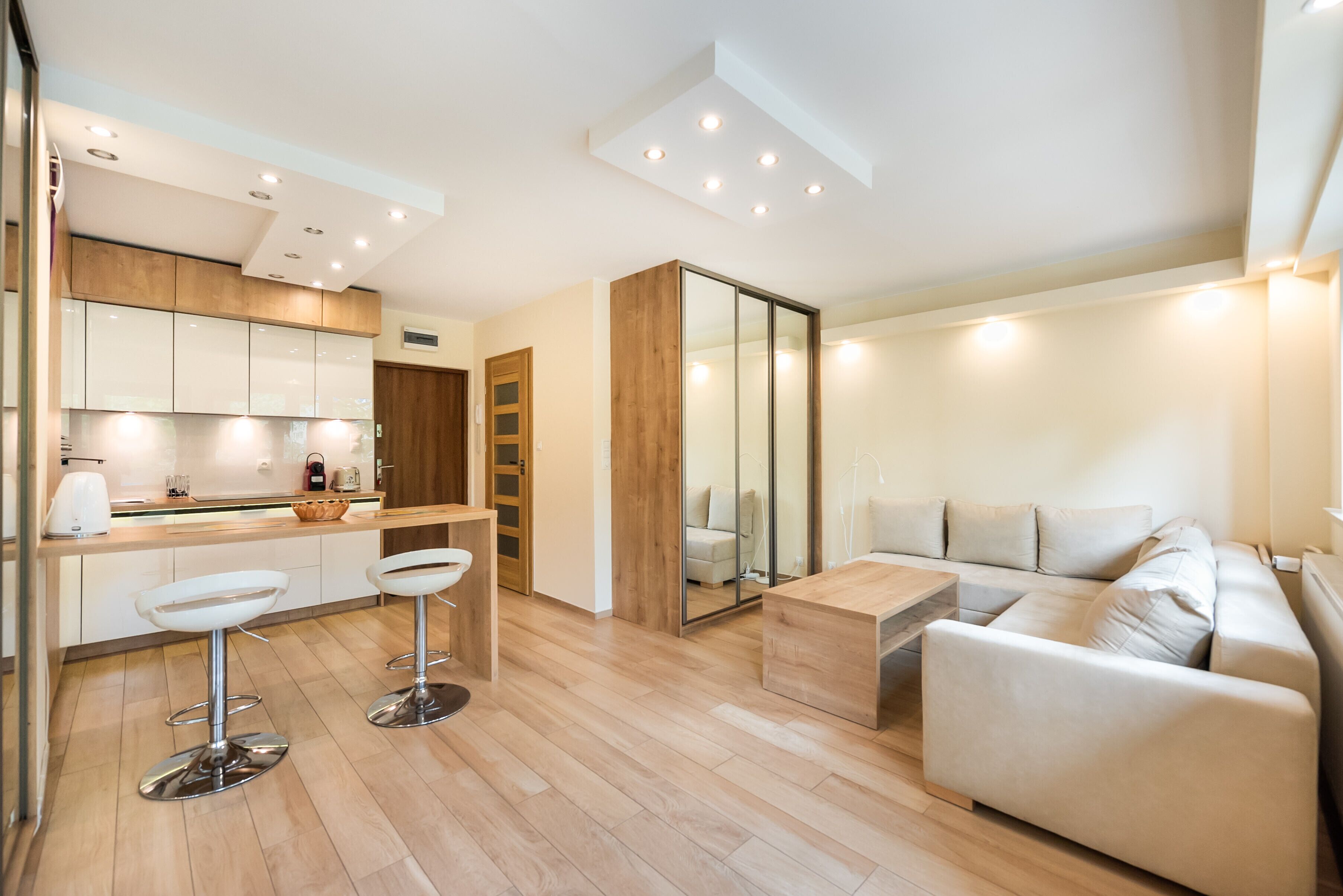 Eine moderne Küche mit weißen Schränken, einer Holzarbeitsplatte und zwei Barhocker. Im Wohnbereich steht ein Holztisch mit einer Bank, ein Sofa und eine Glastrennwand.