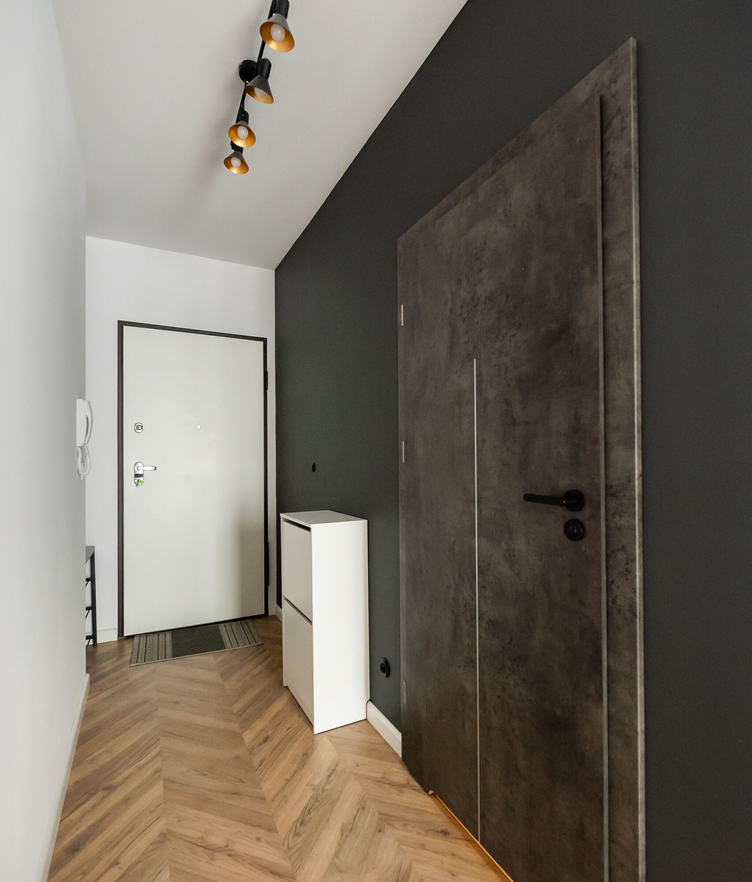 Un couloir avec un parquet en bois disposé en chevrons, une porte blanche, une armoire gris foncé et un petit meuble blanc.