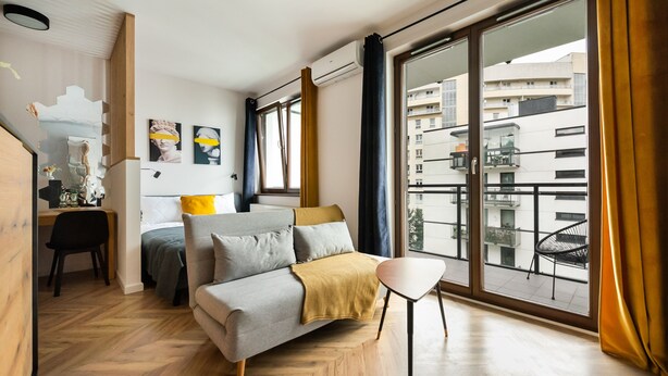 A modern bedroom with a sofa bed, a small desk, and a balcony with a view of the city. - Woronicza 80 Modern Studio (Warszawa)