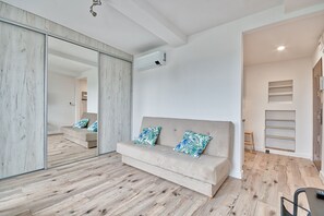 A modern living room with a beige sofa, wooden flooring, a large mirror, and a sliding door leading to another room.