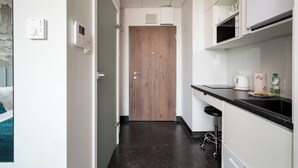 Um corredor estreito com uma porta de madeira no final, uma kitchenette com bancada preta e uma geladeira branca.
