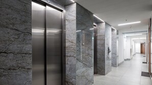 Um corredor moderno com paredes de mármore e chão em mosaico, que dá acesso a elevadores e a outro corredor.