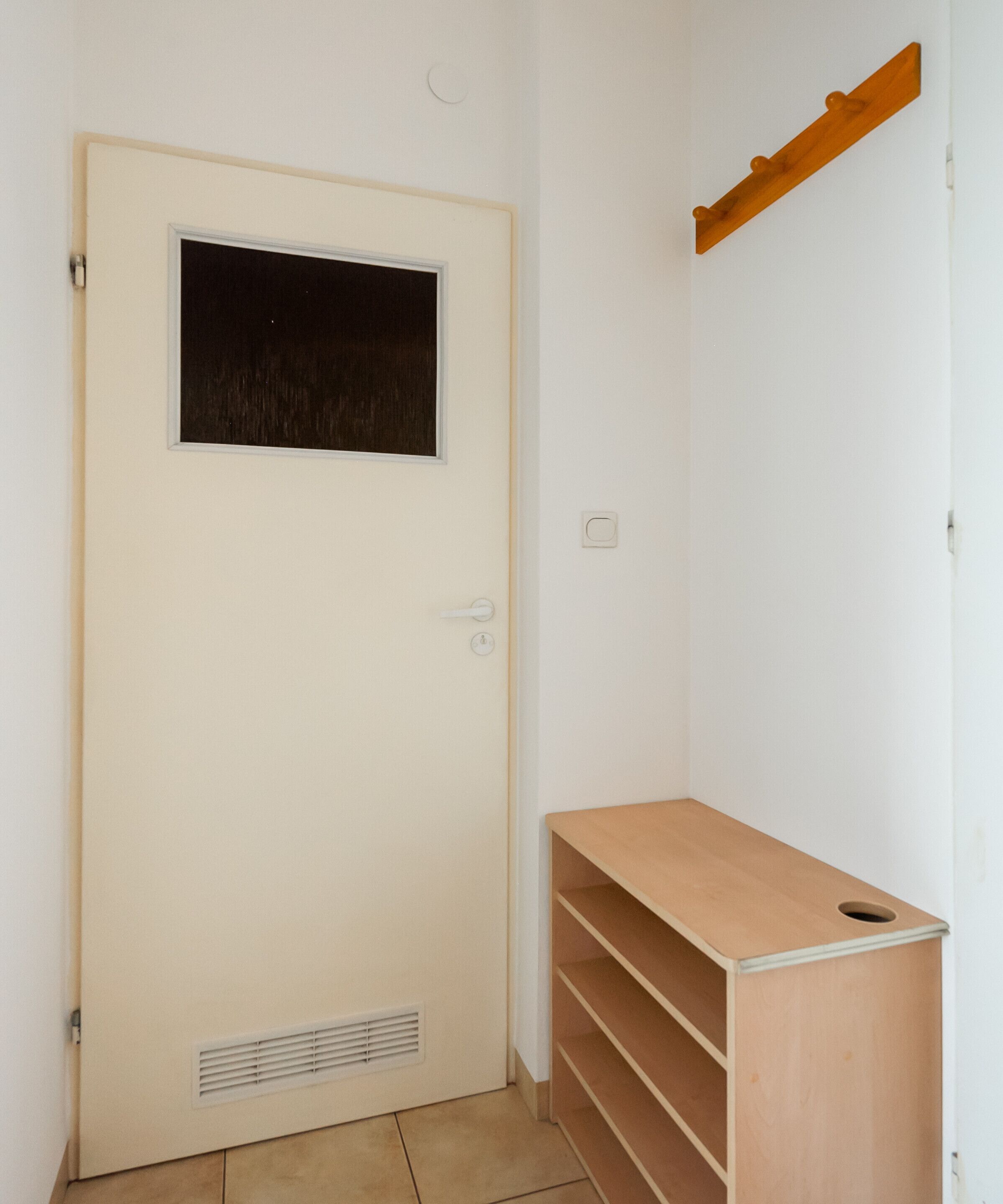 A room with a beige door, a small window, a wooden shelf unit, and a vent.