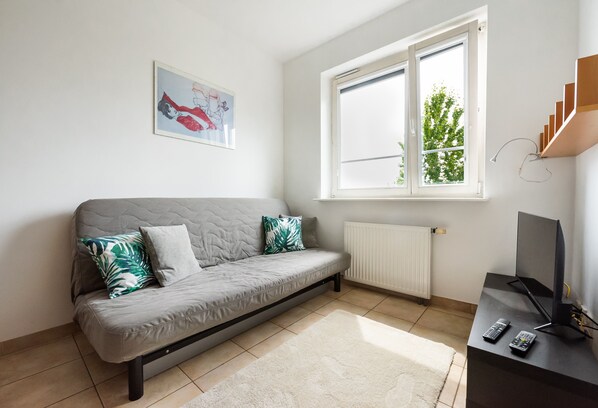 A modern living room with a grey sofa, a white radiator, a window with a view of greenery, and a TV stand with remote controls.