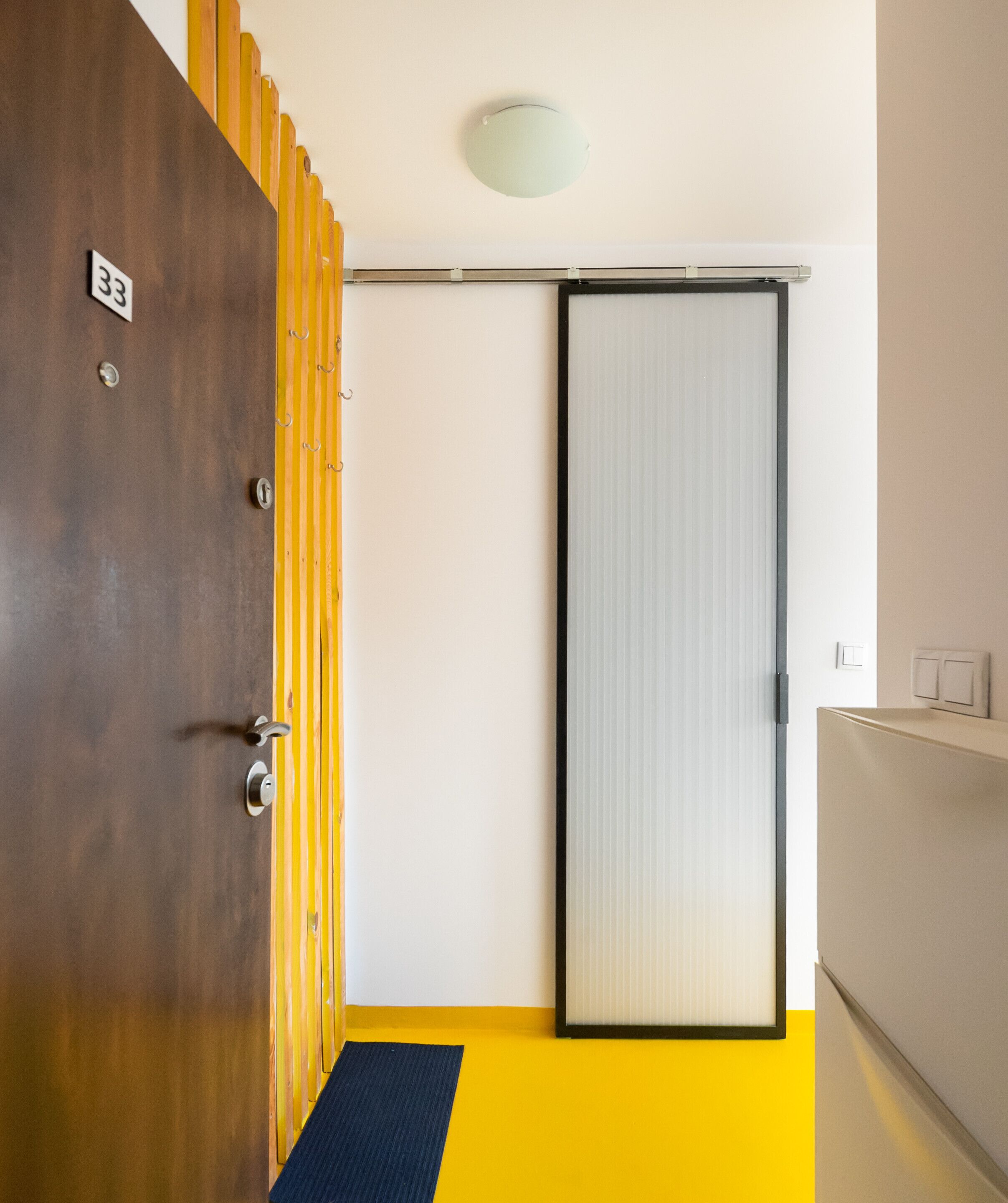 Habitación de hotel con puerta de madera marcada con el número '33', puerta corredera de vidrio, piso amarillo y un tapete azul.