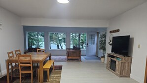 Un salon spacieux avec une table à manger en bois, des chaises, un banc, un téléviseur et une grande fenêtre donnant sur les arbres.