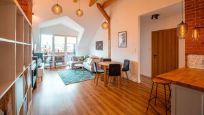 Amplia sala de estar con techos altos, vigas de madera, una mesa de comedor con sillas, una zona de cocina y un gran ventanal que proporciona luz natural.