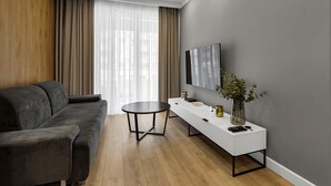 A modern living room with a grey sofa, a round coffee table, a white console table with a TV, and a large window with sheer curtains. - Lotnisko Chopina Modern Apartment (Warszawa)