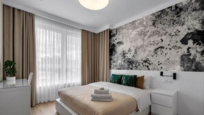 A modern bedroom with a large bed, a bedside table, a nightstand, a wall-mounted light, and a window with sheer curtains. - Lotnisko Chopina Modern Apartment (Warszawa)