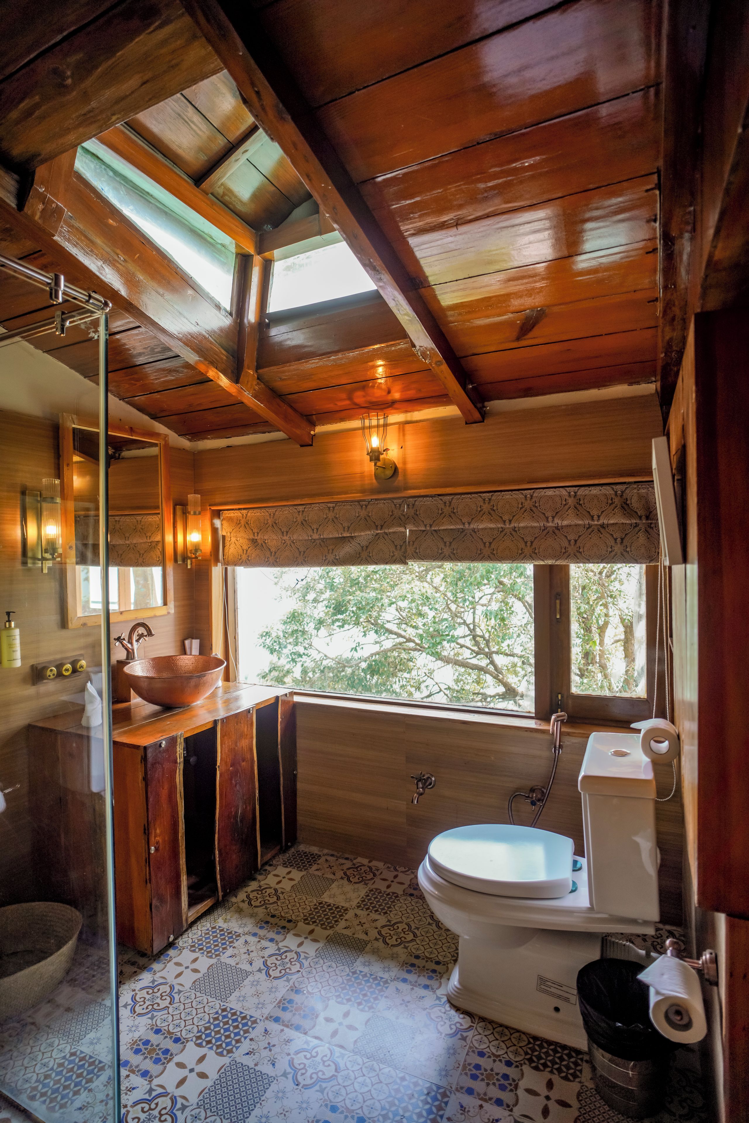 Een badkamer met een houten plafond, een groot raam, een koperen wastafel en een toilet.