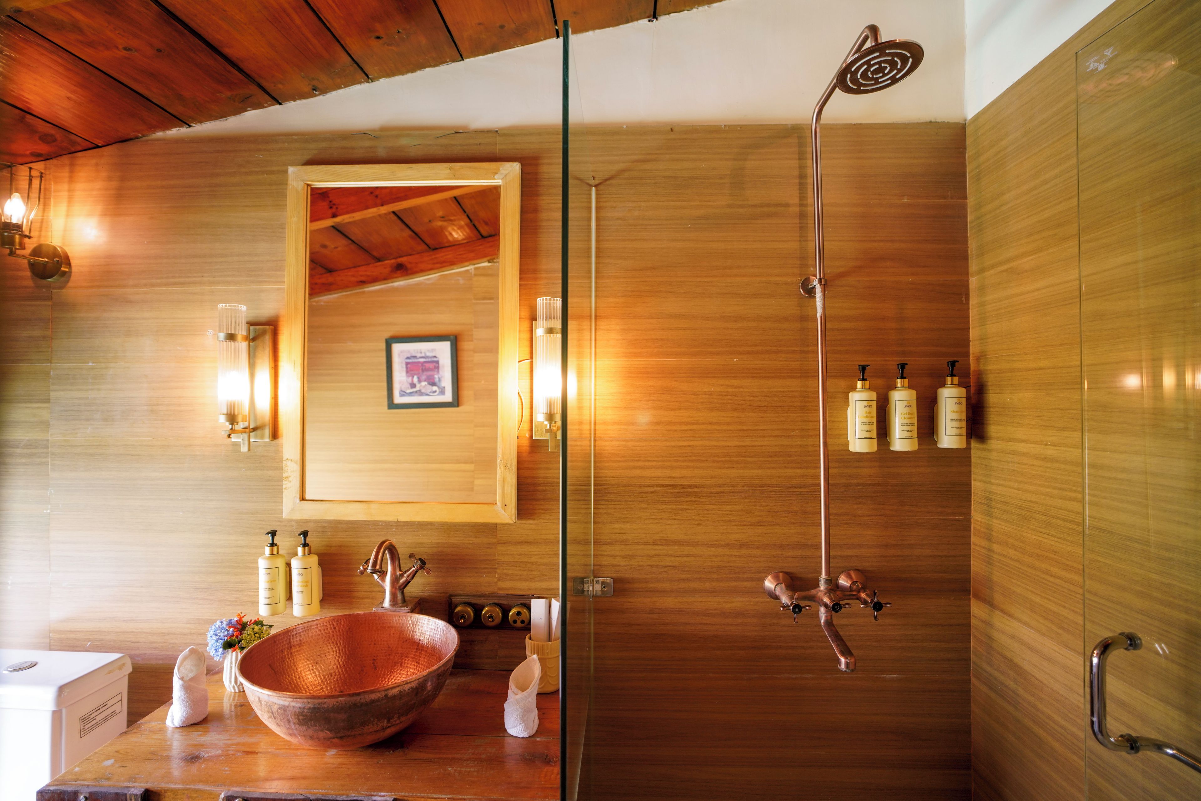 A bathroom with a wooden interior, a large copper basin, a shower with a rain showerhead, and toiletries on the counter.