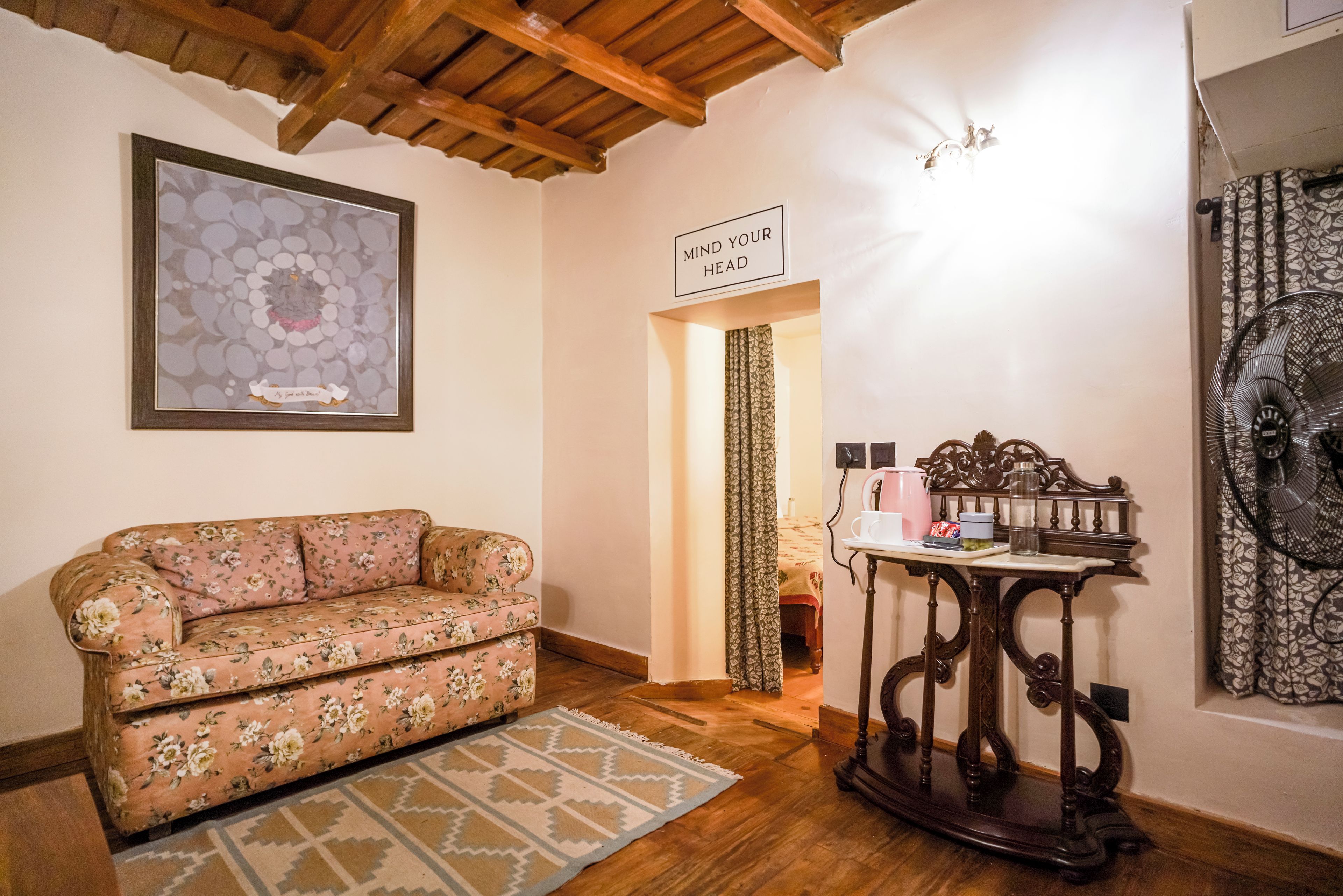 A room with a floral sofa, a wooden table with a kettle, and a sign that says 'MIND YOUR HEAD'.