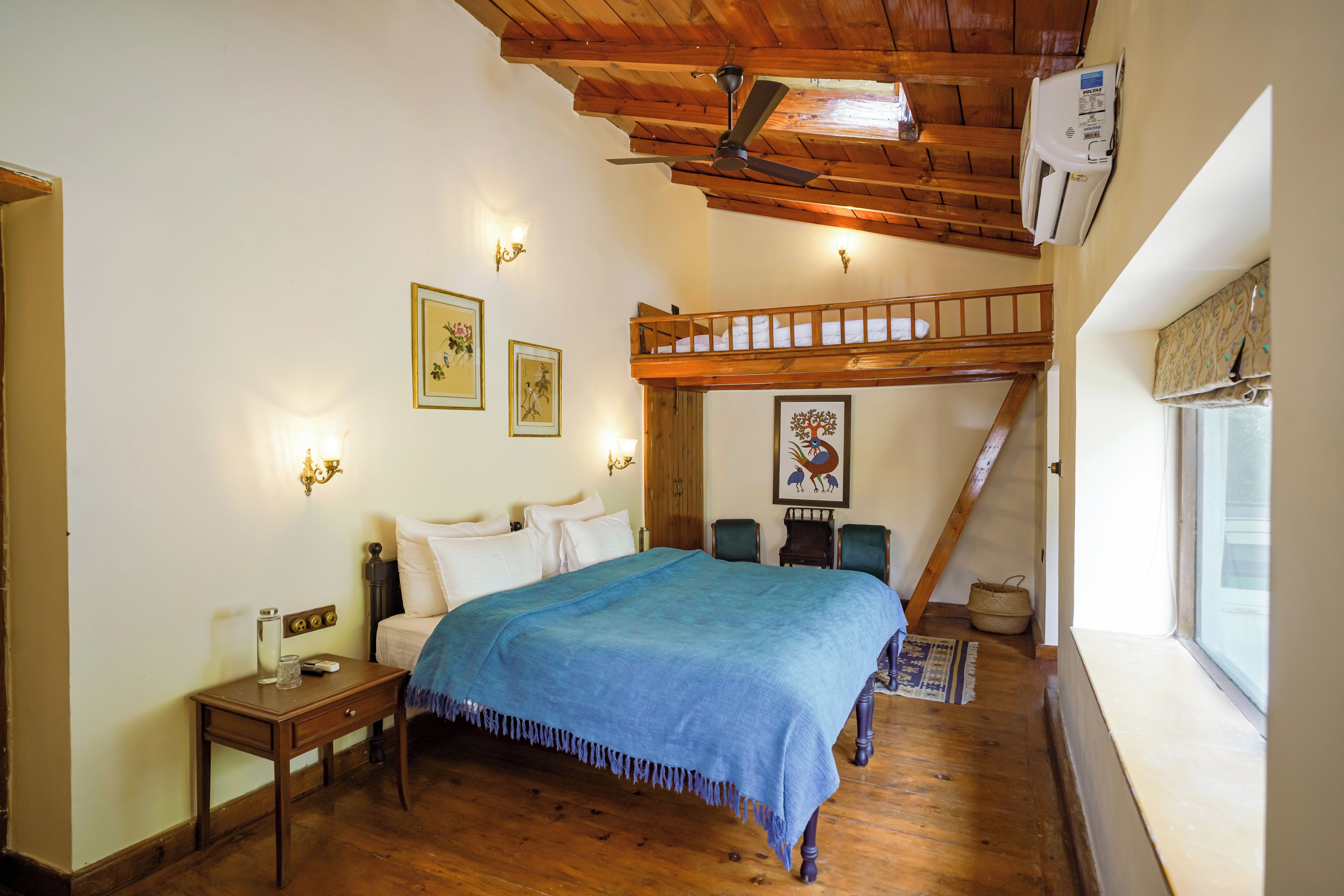 A bedroom with a bunk bed, wooden flooring, a bed with a blue blanket, bedside tables, wall-mounted lights, and framed artwork.