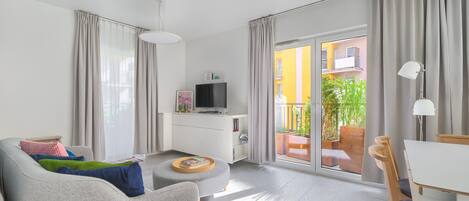 A modern living room with a sofa, ottoman, and a dining area with a table and chairs. A TV is mounted on a cabinet, and there is a sliding glass door leading to an outdoor area with plants.