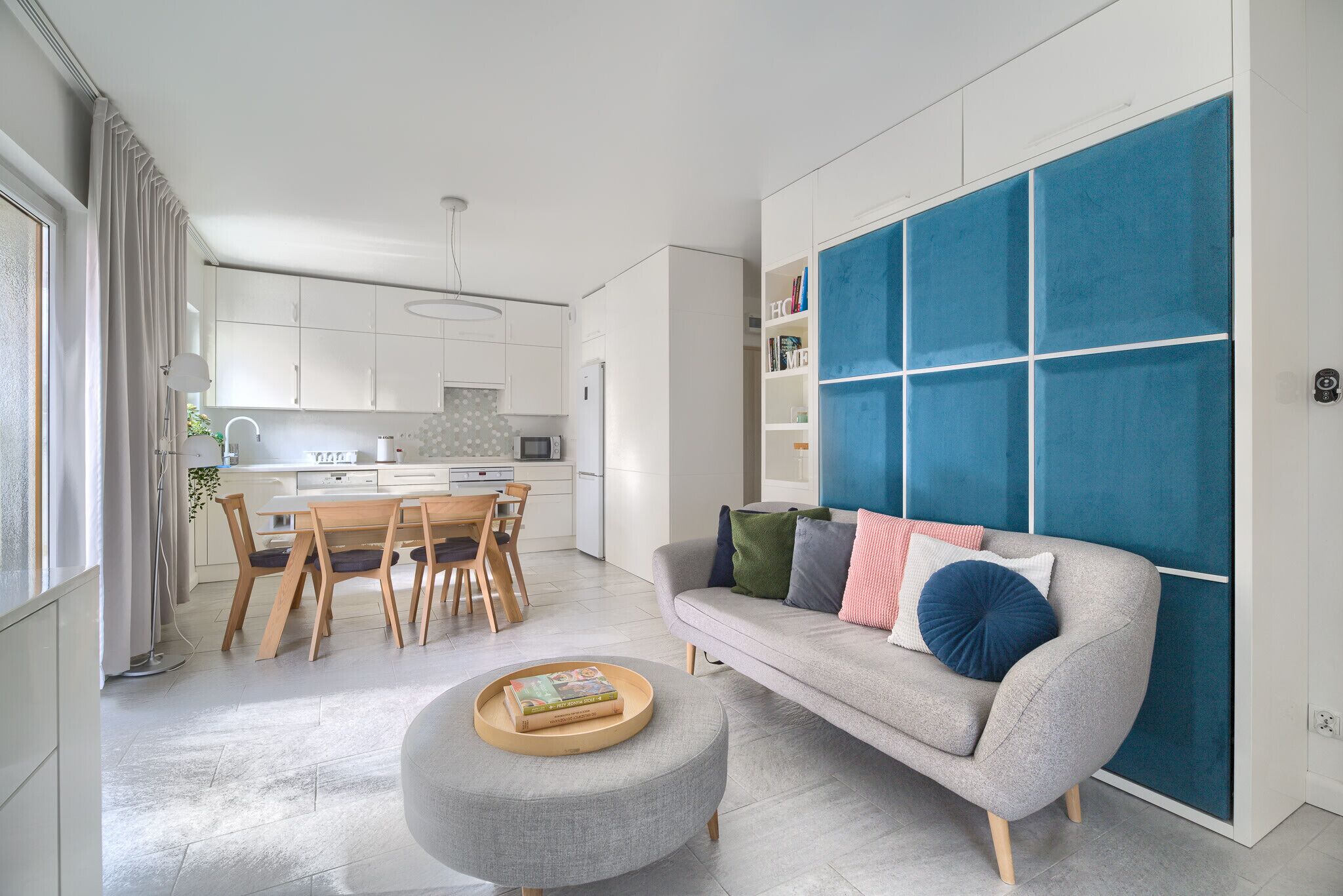 Een moderne woonkamer met een grijze bank, een rond voetenbankje en een eethoek met een houten tafel en stoelen.