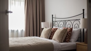 A neatly made bed with a white comforter and several pillows, a bedside table with a lamp, and a window with curtains.
