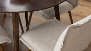 A round wooden table with a bowl of cereal, surrounded by chairs with light fabric upholstery.