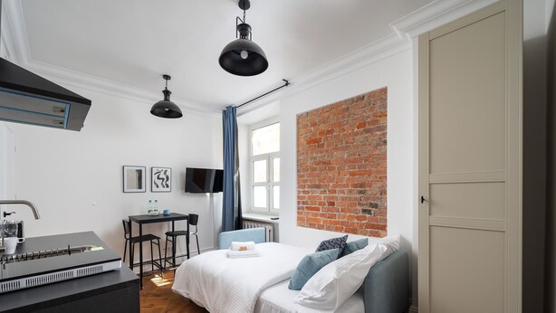 A modern bedroom with a white bed, blue pillows, a brick accent wall, a desk with a computer, and a TV. - Metro Wileński Varsovia Studio #2 (Warszawa)