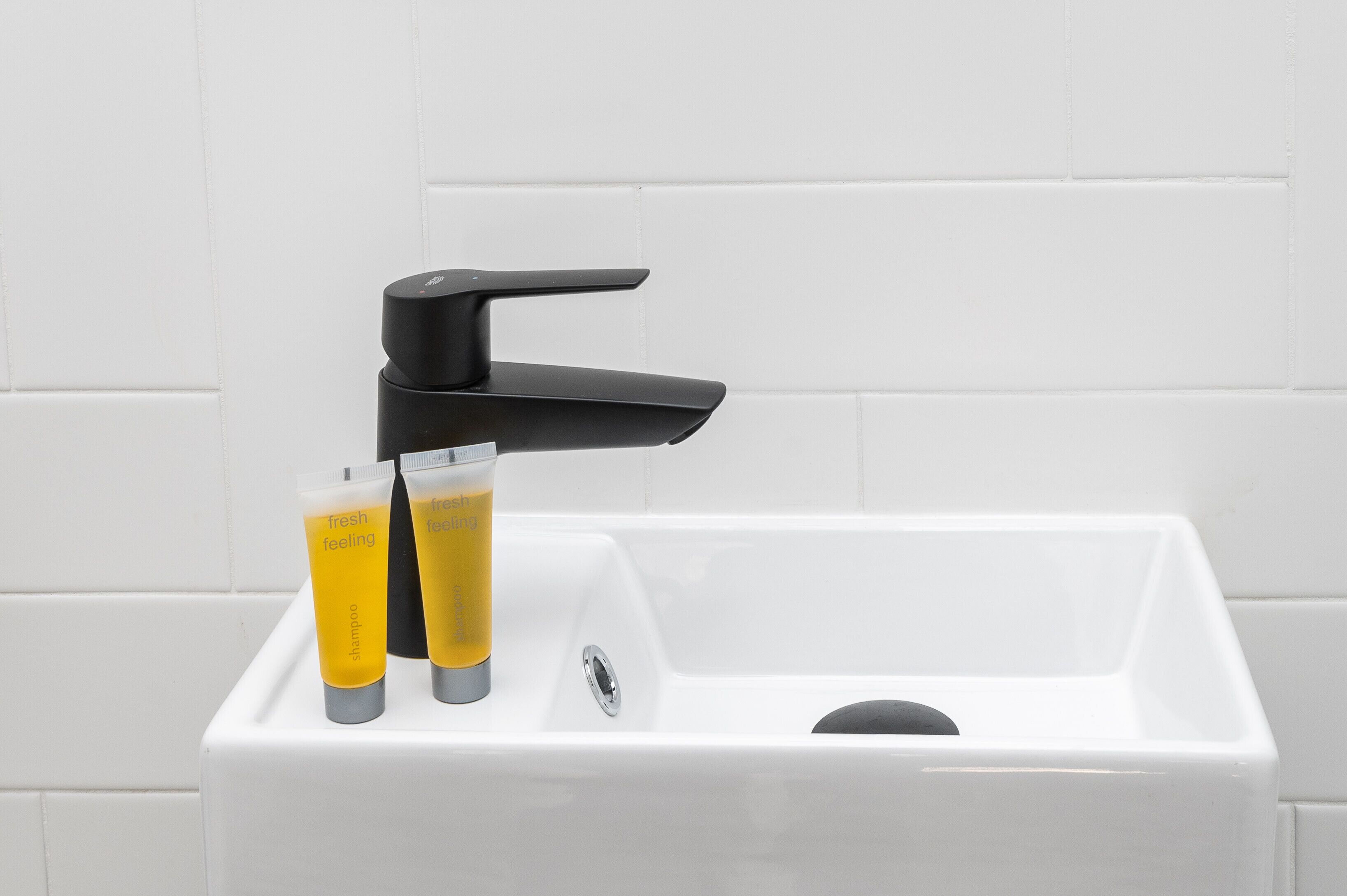 A white sink with a black faucet and two yellow tubes labeled "Fresh Feeling" on a white tiled wall.