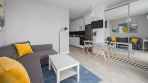 Ett modernt vardagsrum med en grå soffa, gula kuddar, ett vitt soffbord och en matplats med trästolar och ett litet bord.