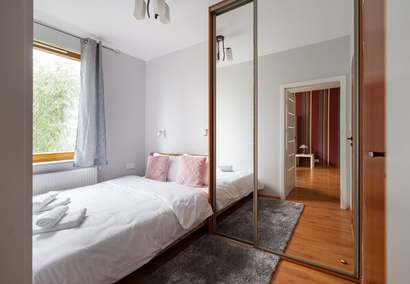 A bedroom with a bed, a mirror, a window with curtains, a radiator, and a hallway with a door.