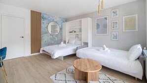Een moderne hotelkamer met twee enkele bedden, een ronde houten salontafel, een blauwe fauteuil en een grote spiegel aan de muur.
