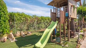 Ein hölzernes Spielhäuschen mit Rutsche, umgeben von einem eingezäunten Garten mit verschiedenen Pflanzen und Steinen.