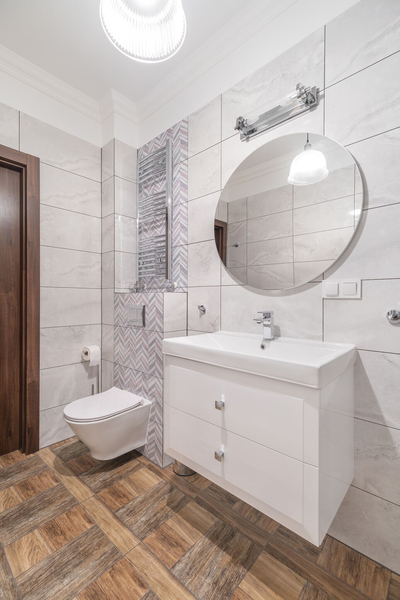 Une salle de bain moderne avec un meuble-lavabo blanc, un miroir rond, des toilettes et un pare-feu en motif chevrons.