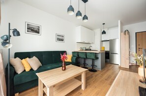 Un salon moderne avec un canapé vert, une table basse en bois et des suspensions noires. On aperçoit, au fond, une cuisine avec des meubles blancs et un îlot central en bois.