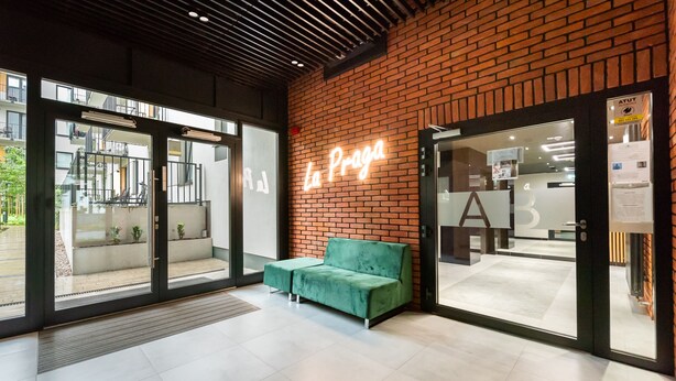 Een modern interieur met een neonbord waarop 'La Praga' staat, een groene bank en een glazen deur naar buiten.