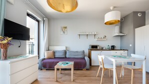 Una habitación moderna y bien iluminada con sofá, una mesa de comedor con sillas, un televisor montado en la pared y una zona de cocina con microondas y cafetera.