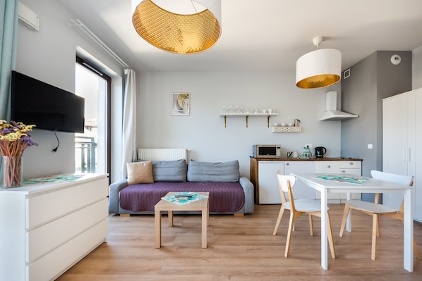 A modern, well-lit room with a sofa, a dining table with chairs, a TV mounted on the wall, and a kitchen area with a microwave and a coffee maker.