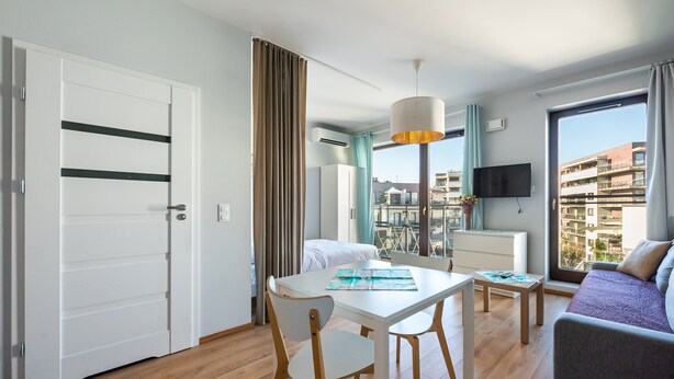 Habitación de hotel moderna con zona de comedor, sofá, televisión y balcón con vista a la ciudad.